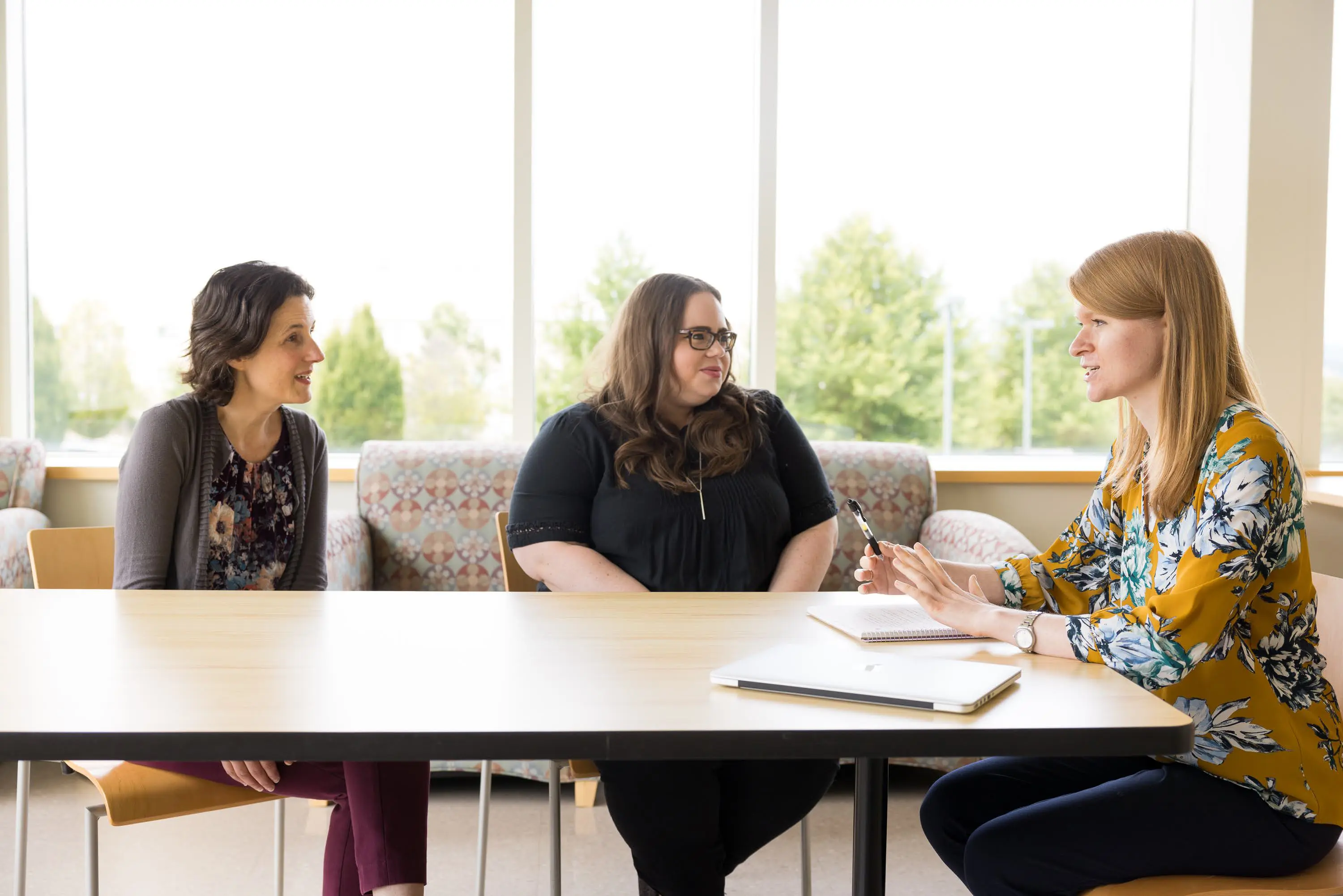 A liaison librarian gives a consultation to two people at a table in room with large windows.